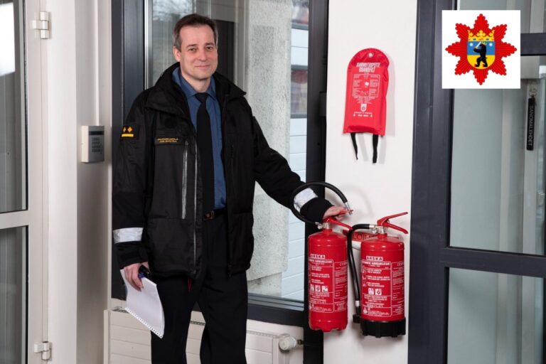Kuvassa palotarkastaja seisoo uniformu päällä pelastuslaitoksen sisääntuloaulassa. Hän pitää vasenta kättään seinällä olevan käsisammuttimen päällä. Toisessa kädessä hänellä on muistiinpanovälineet.
