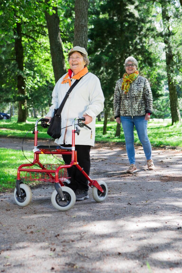Vanhus rollaattorin kanssa kesäisessä maisemassa, taustalla seuralainen