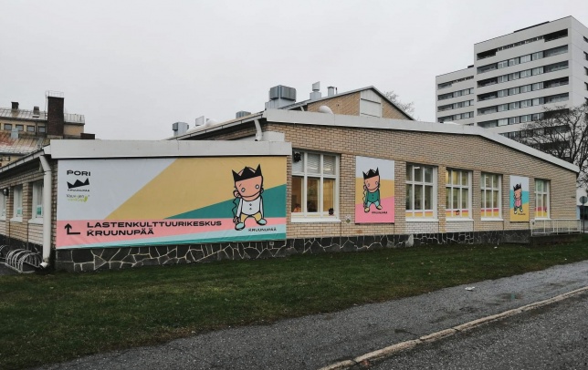Julkisivukuva lastenkulttuurikeskuksesta, edessä katu, nurmikkoalue ja rakennuksen seinissä paljon graafisia kuvia ja ikkunateippauksia.