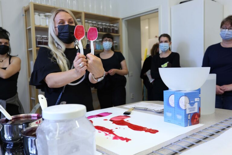 Sisätila, henkilöitä pöydän ympärillä. Luennoivalla henkilöllä kädessään kaksi kauhaa, missä kahta eri väristä maaliainetta.