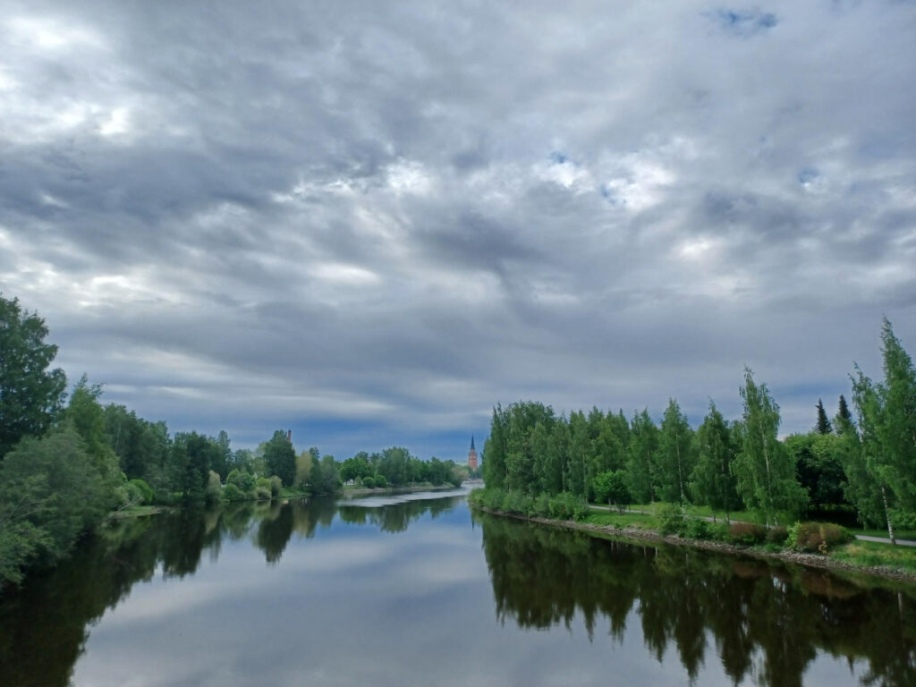jokinäkymä jossa sinivalkoinen taivas heijastuu veteen ja molemmilla puolilla viherpenkat