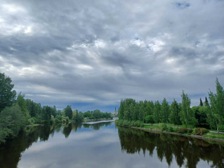 jokinäkymä jossa sinivalkoinen taivas heijastuu veteen ja molemmilla puolilla viherpenkat
