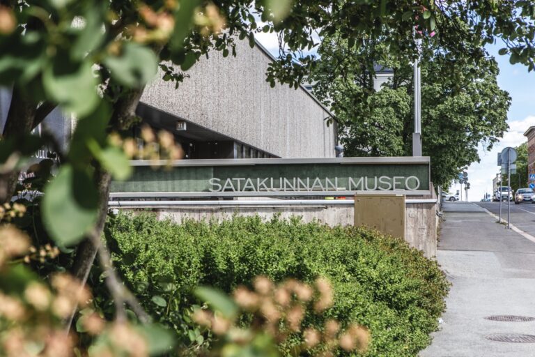 betoninen, kadun varressa sijaitseva rakennus siintää vasemmalla pensaiden ja kukkien takana