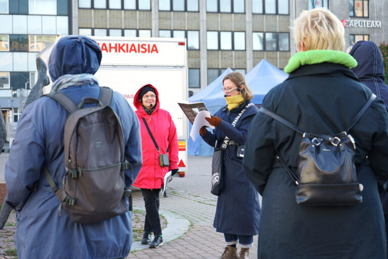 Ryhmä seisoo Porin torilla. Taustalla näkyy ruskea korkea rakennus. Keskellä kasvot kuvaan päin seisoo puna- ja sinitakkiset naiset. Kuvan etualalla selin kaksi sinitakkista ja reppuselkäistä hahmoa.
