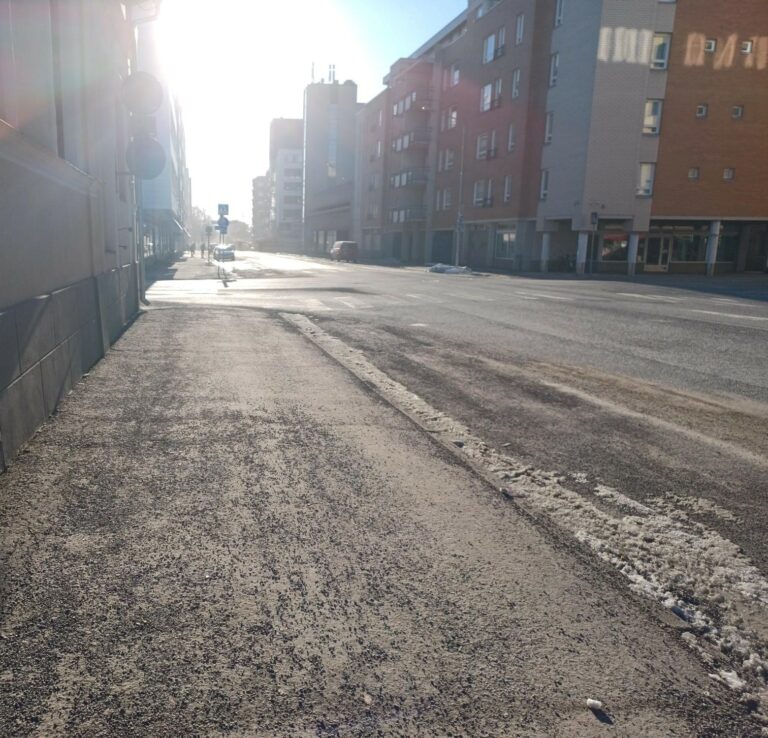 Kuva kadusta jossa paljon hiekoitusta, taustalla kerrostalo ja auringonpaiste.