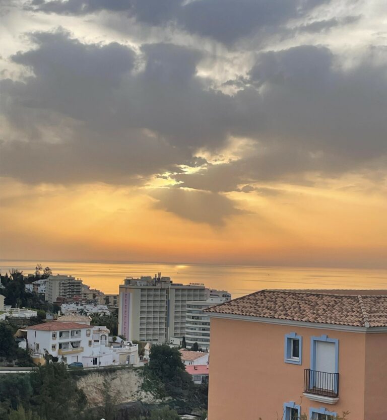 Taustalla punertava aurinko laskemassa mereen, alareunassa kerrostaloja