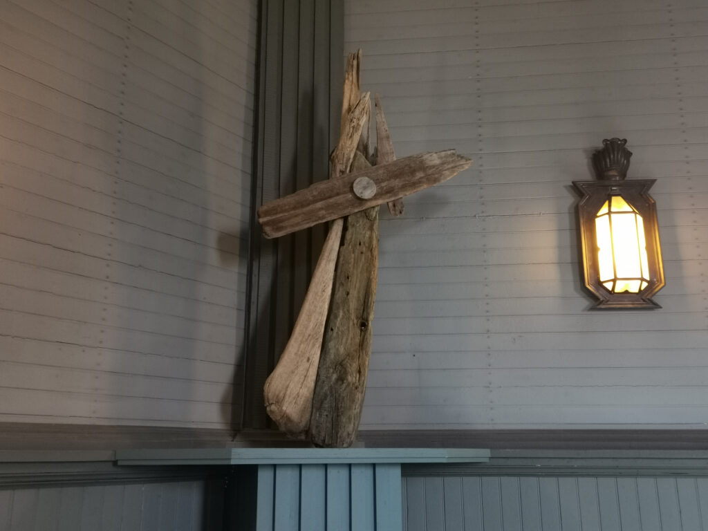 Puinen ristiä muistuttava veistos vaalean puuseinän edessä