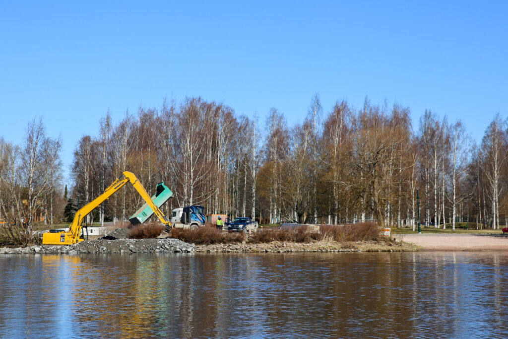 Kirjurinluodon rannan vieressä oleva kaivuupaikka, jossa autoja ja kaivuri. Joki etualalla, takana Kirjurin puita.