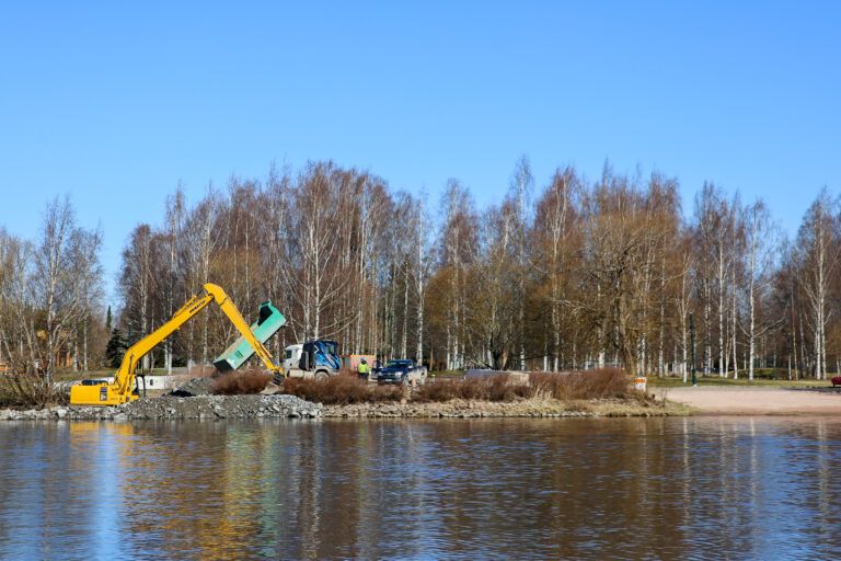 Kirjurinluodon rannan vieressä oleva kaivuupaikka, jossa autoja ja kaivuri. Joki etualalla, takana Kirjurin puita.