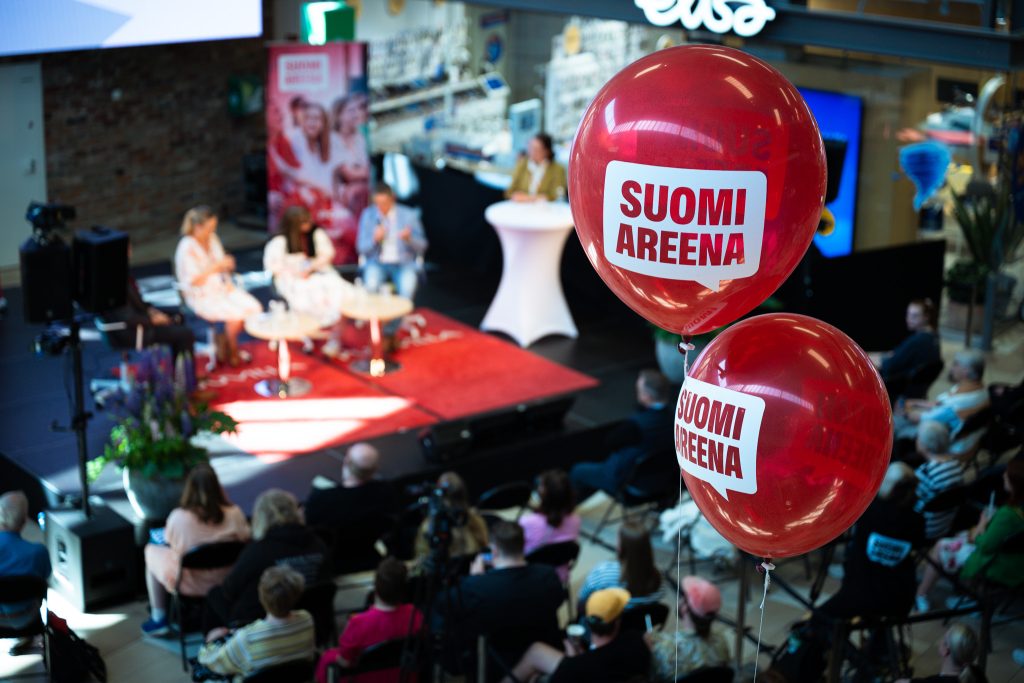 Kaksi punaista SuomiAreena-ilmapalloa lähikuvissa ja taustalla Puuvillan ostoskeskuksen lava jossa puhujia ja ihmisiä yleisössä