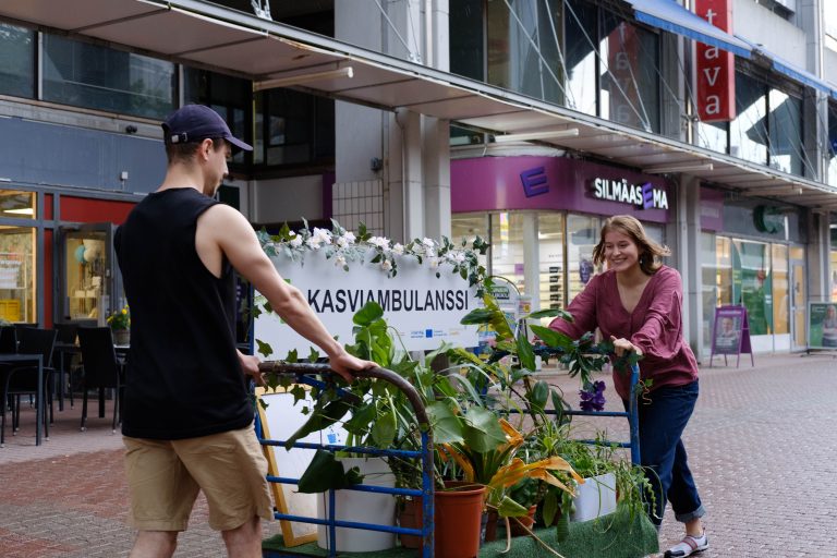 Kuvassa kaksi henkilöä työntää Kasviambulanssia kadulla, kyydissä useita huonekasveja.