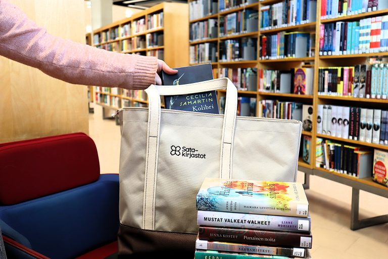 Tuolilla kirjaston kirjahyllyjen edessä kangaskassi, jossa lukee Satakirjastot ja edessä on kasa kirjoja.
