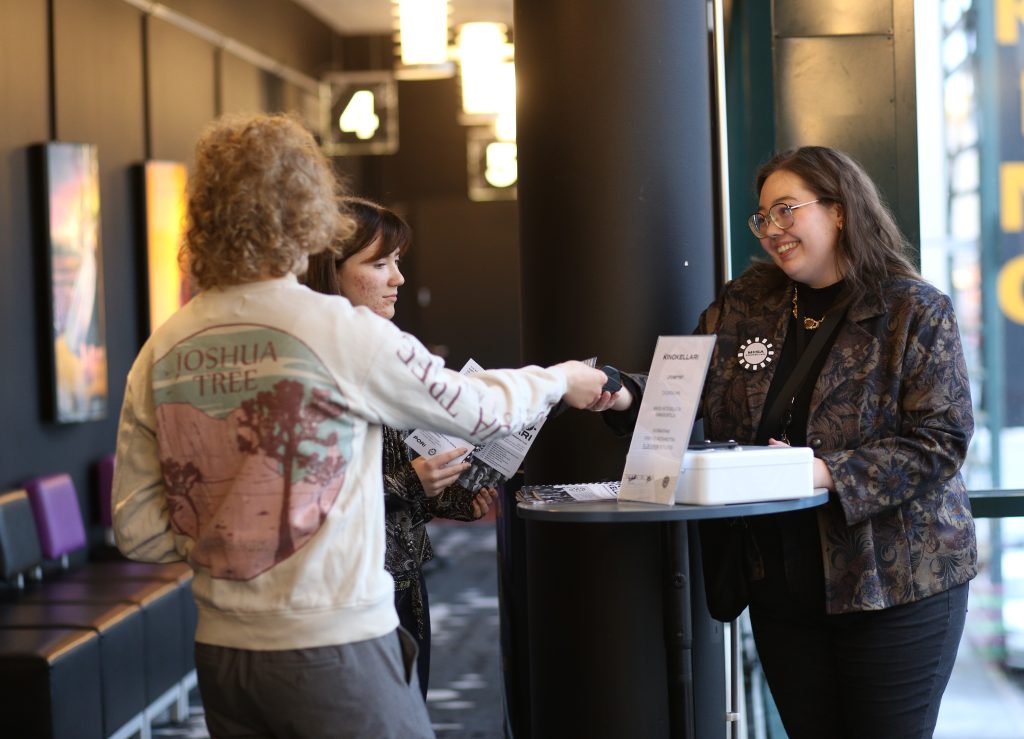 Kinokellarin myyntipiste. Kinokellarin vastaava hymyilee ja ojentaa maksupäätettä kahdelle nuorelle aikuiselle. Kaksi nuorta aikuista ovat nainen ja mies. Miehen kasvot eivät näy. Myyntipiste sijaitsee Porin Promenadikeskuksen toisessa kerroksessa.