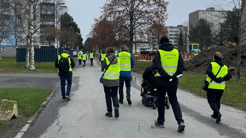 Joukko ihmisiä keltaiset liivit päällä kävelemässä kerrostalojen keskellä.