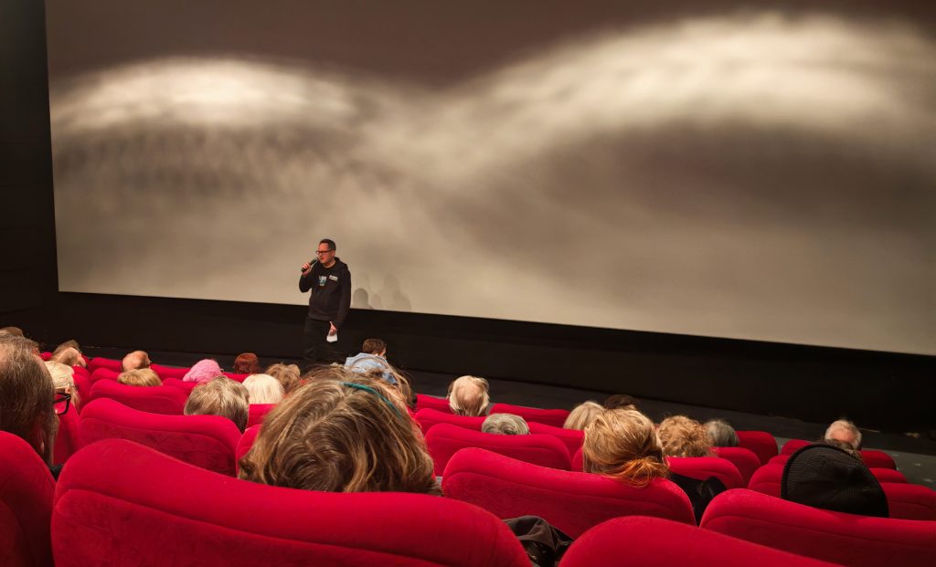 Mies puhumassa elokuvateatterin valkokankaan edessä. Kuvassa etualalla punaisia istuimia, joissa istuu ihmisiä, kuvattuna takaapäin.