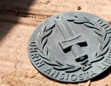 Harmaa, pyöreä muistolaatta puisella pohjalla. Laatassa lukee uhrin ansiosta lippu liehuu.