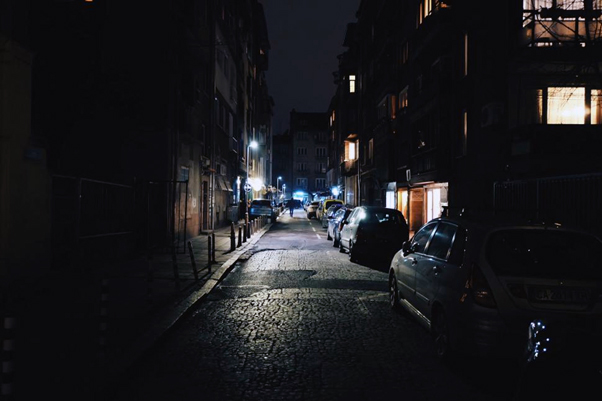 In the picture there is an empty, dimly lit, old city street at night. Only one man is walking far in the distance. There are cars parked on the right side of the street. The ground is rocky.