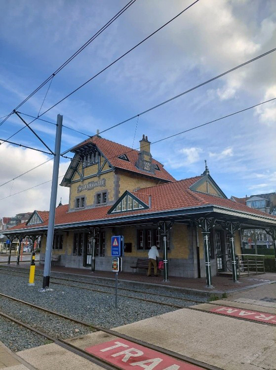 Kuva Belgiassa sijaitsevan Knokken vanhasta, koristeellisesta rautatie- ja raitiovaunuasemasta. Rakennus on tyyliltään perinteinen, ja siinä on punainen tiilikatto, koristeellisia puurakenteita ja keltaisen sävyinen tiiliseinä.