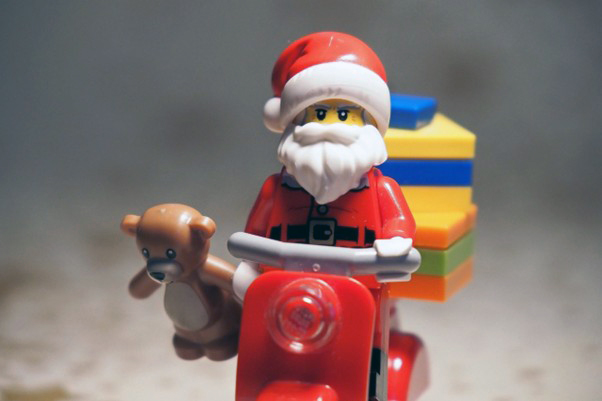 Lego-joulupukki ajaa punaista skootteria samalla pitäen kiinni pienen ruskean nallen kädestä. Pukin takana skootterin penkillä on kasa joululahjoja. Kuva: Pixabay, Ralf 1403
