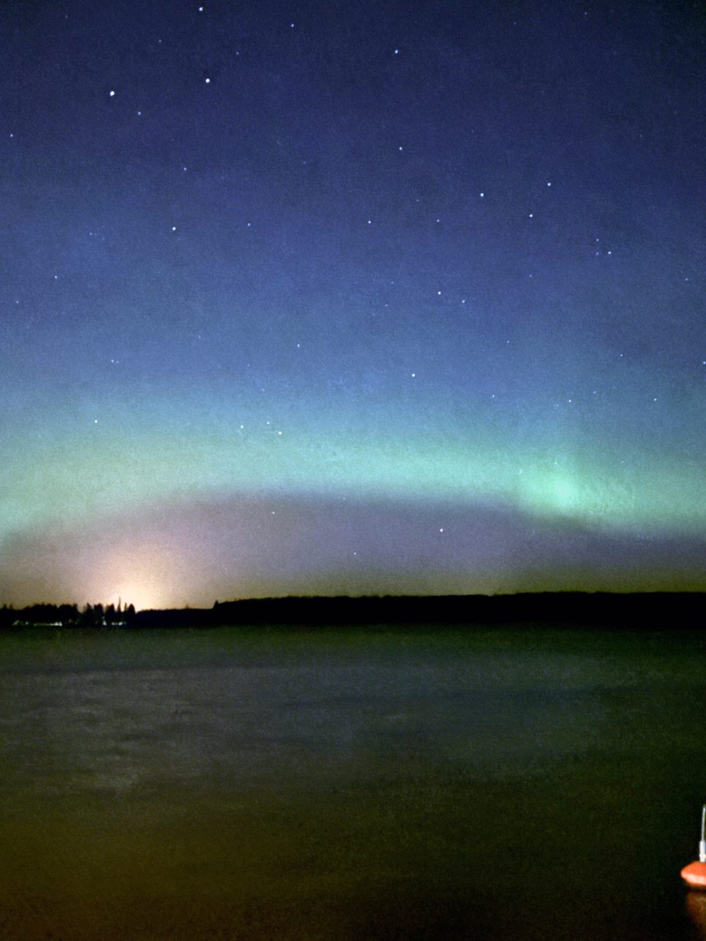 Maisemakuva, jossa näkyy yötaivas ja revontulia. Etualalla on vesistö, joka heijastaa hieman taivaan värejä.