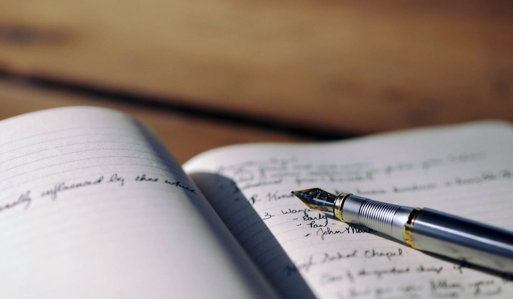 Kuvituskuva, jossa näkyy lähikuva avoimesta muistikirjasta, johon on kirjoitettu käsin mustalla musteella. Muistikirjan päälle on asetettu mustekynä.