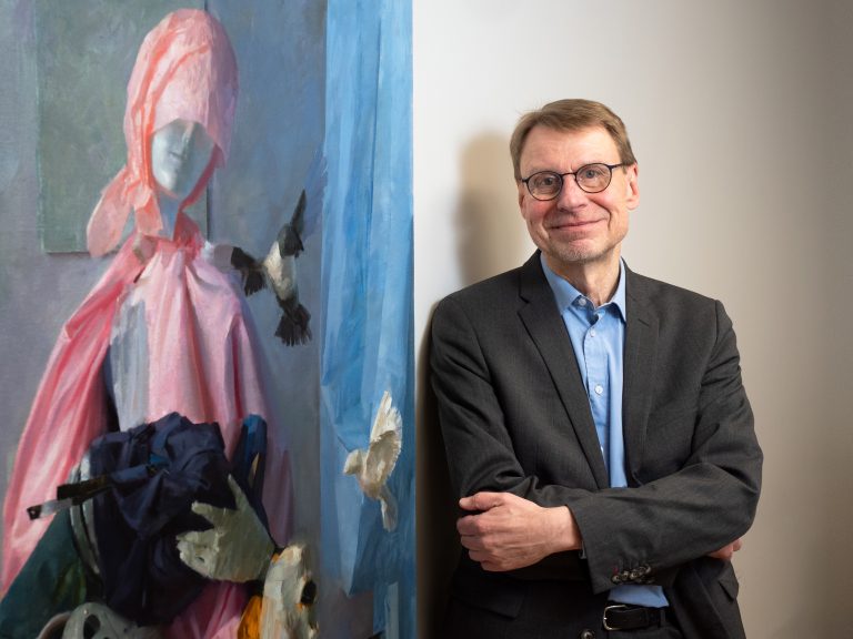 Vasemmalla puolella kuvaa taulu jossa naishahmo. Oikealla puolella kuvaa hymyilevä Arto Jurttila.