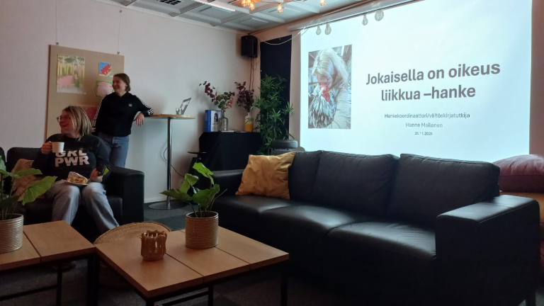 Viihtyisä tila Ystäväkahvila Murussa, jossa kaksi henkilöä sohva-alueella ja seinälle heijastuu esitys ‘Jokaisella on oikeus liikkua -hanke’