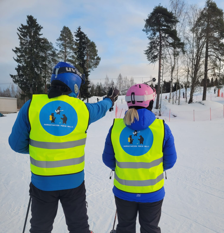 Kaksi huomioliiviin pukeutunutta henkilöä laskettelurinteen juurella, sukset jalassa.