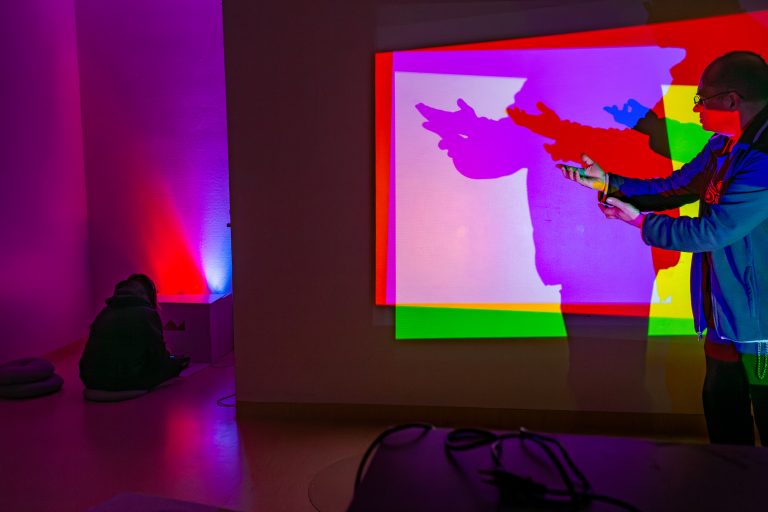 Ihmisiä katselemassa ja leikkimässä seinään heijastuvien värikkäiden valojen kanssa.