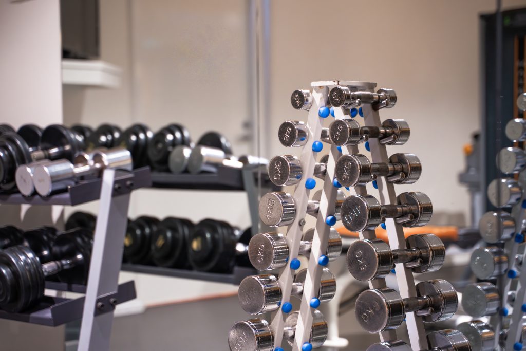 Keskustan uimahallin kuntosalin hyllyissä ja telineissä olevia erikokoisia käsipainoja, osa metallisia ja osa mustia, järjestettyinä riveihin ja pinoihin kirkkaasti valaistussa sisätilassa.
