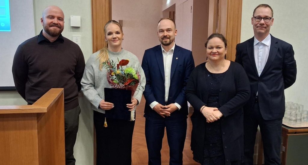 Viisi hymyilevää ihmistä rivissä, yhdellä kädessään kukkakimppu.