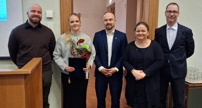 Viisi hymyilevää ihmistä rivissä, yhdellä kädessään kukkakimppu.