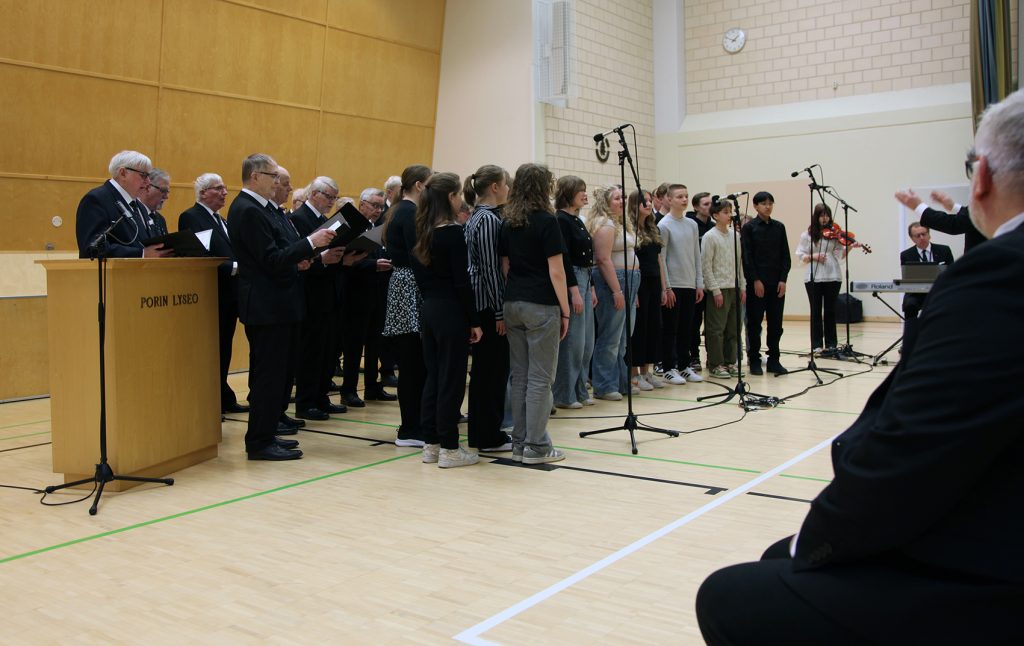 Kuva vuoden 2025 kansallisen veteraanipäivän juhlan musiikkiesityksestä, jossa Porin Lyseon koulun musiikkiluokan oppilaat ja Perinnekuoro Vaarinlaulu esiintyvät yhdessä.
