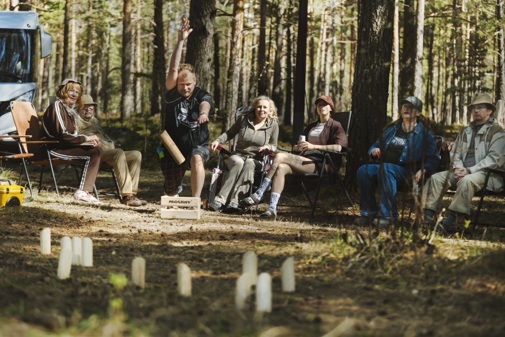 Kuva metsästä, jossa etualalla mölkkypalikoita pystyssä, taustalla ihmisiä istumassa retkituoleissa.