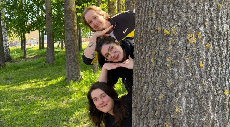 Puunrungon takaa kurkkaamassa kolme hymyilevää etsivää nuorisotyöntekijää.