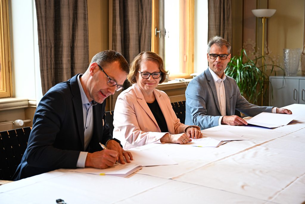 Kolme henkilöä pöydän ääressä allekirjoittamassa sopimuksia