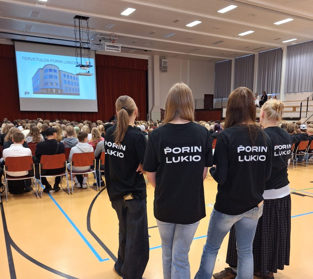 Kuvassa Porin lukion tutoroppilaita seisoo lukion juhlasalin takaosassa. Salin etuosassa istuu suuri joukko ihmisiä riveittäin tuoleilla kohti esiintymislavaa. Valkokankaalla näkyy esitysdia, jossa lukee "TERVETULOA PORIN LUKIOON”.