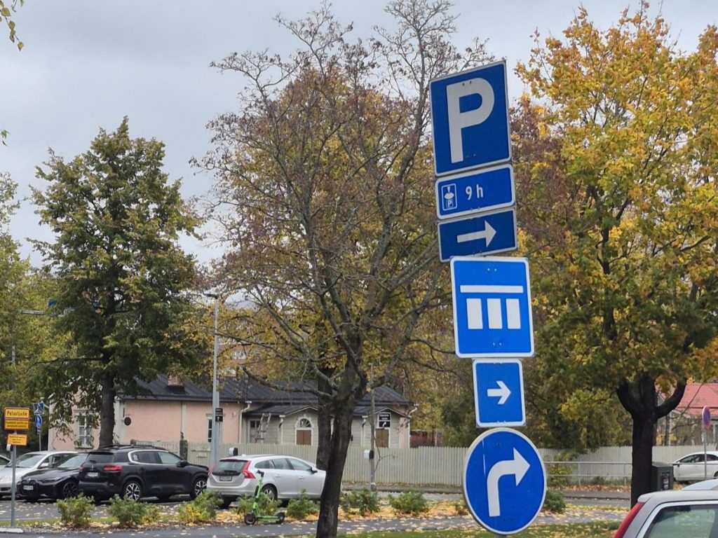 Kuvassa sinivalkoisia pysäköintialueen merkkejä, taustalla syksyisiä puita sekä autoja.